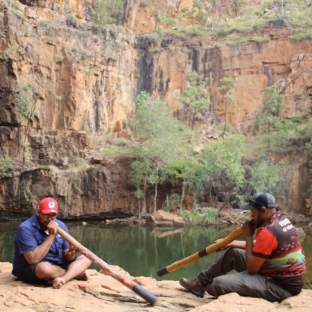 Two people next to water hole playing didgeridoo 
